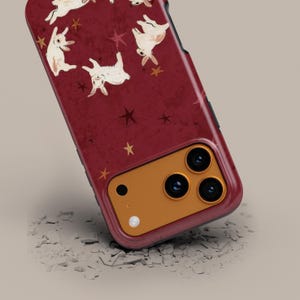 May include: A burgundy phone case featuring a pattern of white rabbits and gold stars. The case has a protective bumper and a gold camera housing. The design is whimsical and suitable for a variety of phone models.