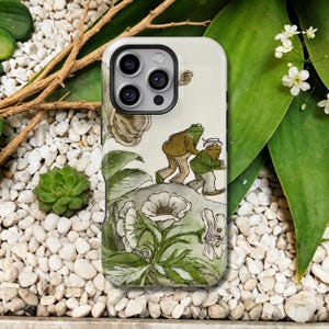 May include: A phone case featuring a whimsical illustration of two frog characters in a vintage style. The design includes a light green background with white flowers and foliage, and a detailed image of the frogs in brown and green attire.