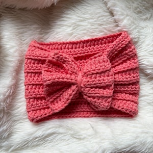 Puede incluir: Una diadema de ganchillo hecha a mano, de color coral, con un gran lazo. La diadema está hecha de un hilo grueso, con un diseño texturizado y acanalado. El lazo está centrado en la diadema.