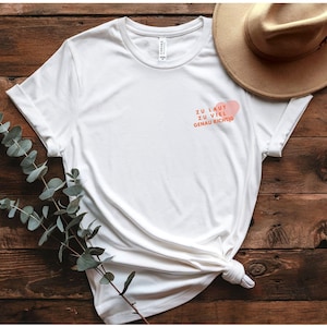 Könnte beinhalten: Weißes T-Shirt mit Rundhalsausschnitt und hochgekrempelten Ärmeln. Das Shirt hat eine kleine rosa Herzgrafik mit dem deutschen Text "ZU LAUT ZU VIEL GENAU RICHTIG" in Orange. Ein beiger Hut liegt auf der Holzoberfläche.