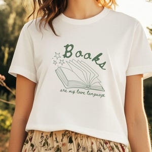 Könnte beinhalten: Weißes T-Shirt mit einer grünen Grafik eines offenen Buches und dem Text "Books are my love language". Das Design enthält Sterne über dem Wort "Books". Das Shirt ist aus weichem Material.