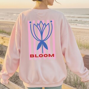 Könnte beinhalten: Ein hellrosa Langarm-Sweatshirt mit einer Grafik auf der Rückseite. Das Design besteht aus vertikalen hellrosa Streifen, einer blauen stilisierten Lotusblumen-Illustration und dem Wort BLOOM in fetten roten Großbuchstaben.