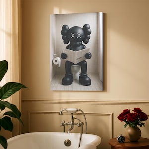 May include: A rectangular wall art print featuring a black cartoon character with X-shaped eyes sitting on a white toilet in a tiled bathroom, reading a newspaper. A white toilet paper roll is mounted on the wall to the left of the character.