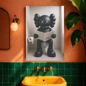 May include: A framed wall art print depicting a matte black KAWS-style character with X-shaped eyes sitting on a white toilet in a tiled bathroom, reading a newspaper. The character has rounded ears and large feet, set against a white tiled wall.