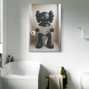 May include: A rectangular wall art print featuring a black KAWS character with X-shaped eyes sitting on a white toilet in a tiled bathroom. The character is holding an open newspaper. A roll of white toilet paper is mounted on the wall to the left.