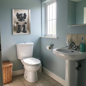 May include: A rectangular wall art print featuring a black KAWS character sitting on a toilet and reading a newspaper. The artwork is displayed in a bathroom with light blue walls, a white toilet, a pedestal sink, and a wicker laundry basket.
