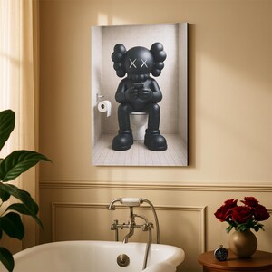 May include: A framed wall art print depicting a matte black KAWS character with X-shaped eyes sitting on a white toilet in a tiled bathroom. The character holds a small black device in its hands, and a roll of white toilet paper hangs on the wall.
