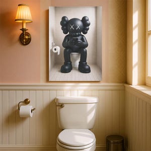 May include: A framed wall art print depicting a black cartoon character with X-shaped eyes sitting on a white toilet while looking at a smartphone. The character is positioned in a tiled bathroom stall with a white toilet paper roll on the wall.