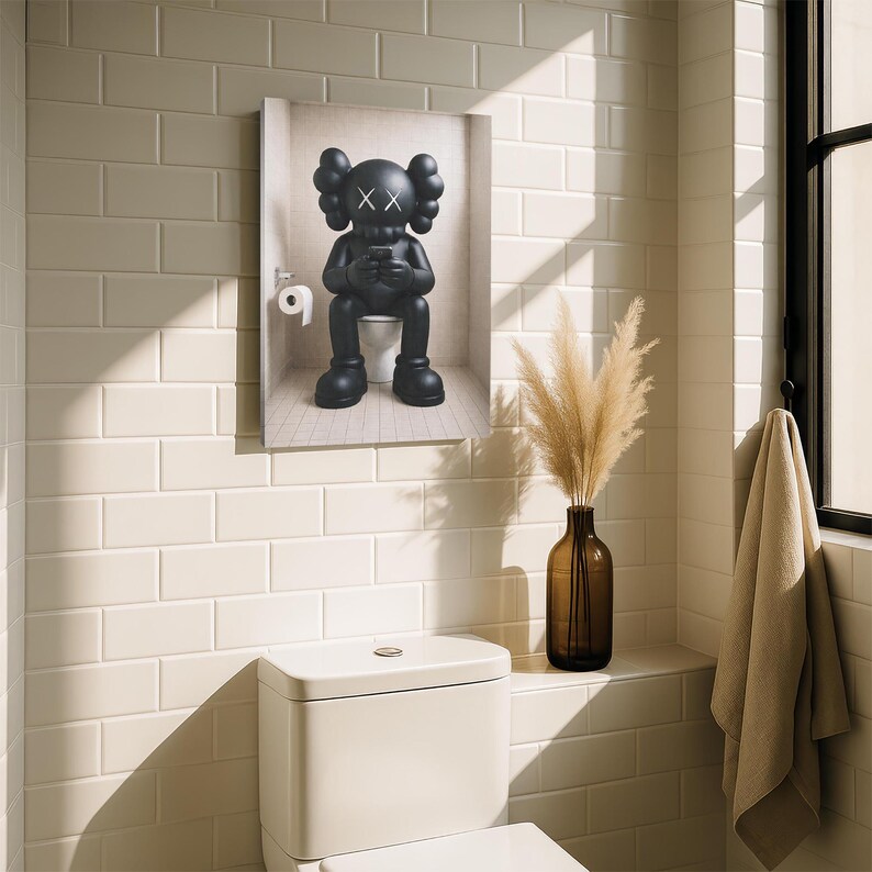 May include: A rectangular wall art print featuring a matte black KAWS character sitting on a white toilet while looking at a smartphone. The character has signature X-shaped eyes and rounded ears. A roll of white toilet paper hangs on the wall nearby.