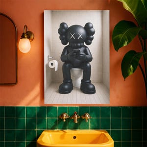 May include: A framed wall art print depicting a matte black KAWS character with X-shaped eyes sitting on a white toilet in a tiled bathroom, holding a smartphone. The character has rounded ears and large feet, set against a white tiled wall.