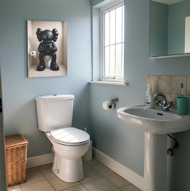 May include: A rectangular wall art print featuring a black KAWS character sitting on a toilet while looking at a smartphone. The character has signature X-shaped eyes and rounded ears. The print is displayed on a light blue bathroom wall.