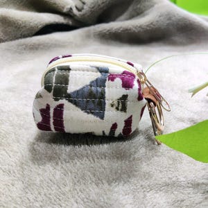 May include: A small, square coin purse with a cream background and a pattern of purple, gray, and green shapes. The purse has a gold zipper and a gold key ring attached. A pink and white flower and green leaf are in the background.