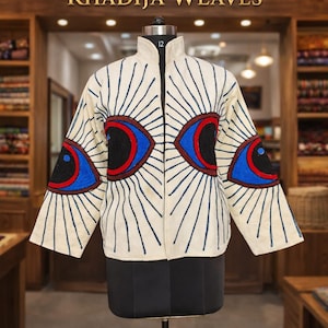 Puede incluir: Chaqueta color crema con cuello alto y cremallera negra. Presenta un diseño único con patrones azules y rojos en forma de ojo en la parte delantera y las mangas, y líneas azules verticales que irradian del cuello. El texto "KHADIJA WEAVES" se muestra arriba.