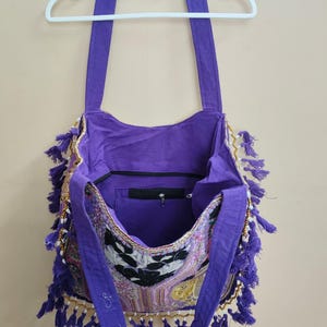 Puede incluir: Un bolso de hombro morado con un panel frontal estampado, adornado con borlas y cuentas doradas. El bolso tiene correas largas y un bolsillo interior con cremallera. El bolso est&aacute; colgado de una percha blanca.
