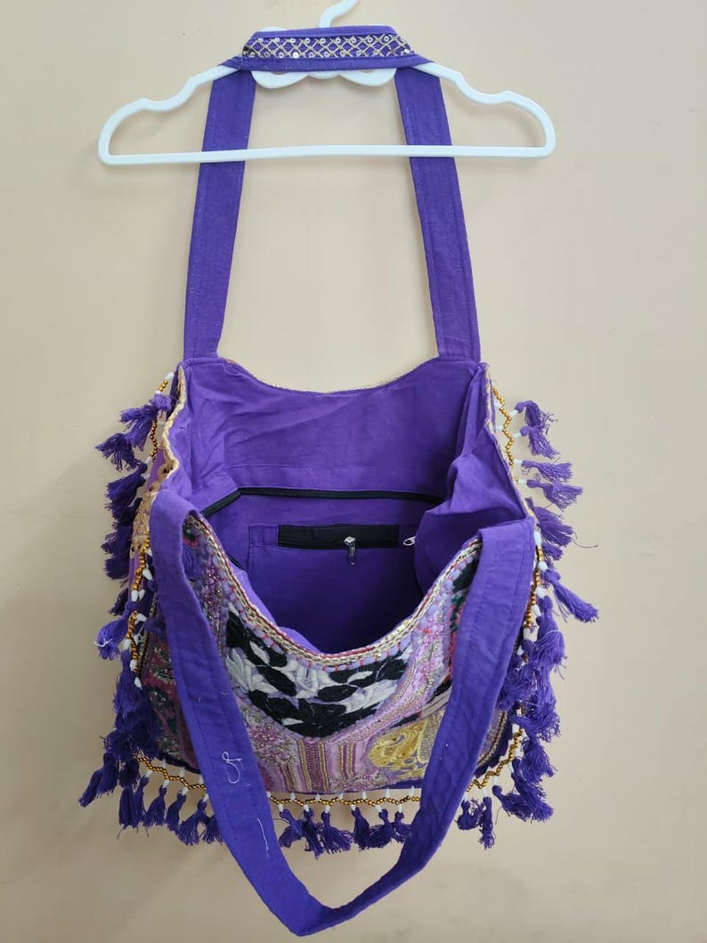 Puede incluir: Un bolso de hombro morado con un panel frontal estampado, adornado con borlas y cuentas doradas. El bolso tiene correas largas y un bolsillo interior con cremallera. El bolso est&aacute; colgado de una percha blanca.
