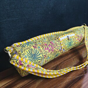 Puede incluir: Una bolsa amarilla para esterilla de yoga con un estampado floral en rosa, azul y verde. La bolsa tiene cierre de cremallera y una correa a juego. La bolsa es de tela y tiene forma rectangular.