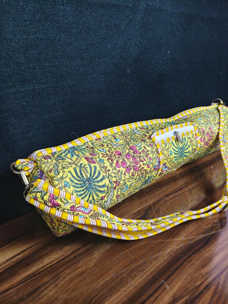 Puede incluir: Una bolsa amarilla para esterilla de yoga con un estampado floral en rosa, azul y verde. La bolsa tiene cierre de cremallera y una correa a juego. La bolsa es de tela y tiene forma rectangular.
