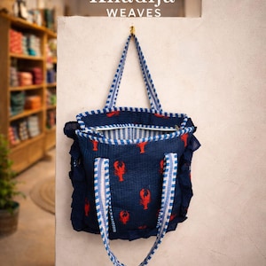 Op de afbeelding: Marineblauwe tote bag met rode kreeftenprint. De tas heeft blauw-wit gestreepte handvatten en randen. De tekst "Khadija WEAVES" staat bovenaan de afbeelding.