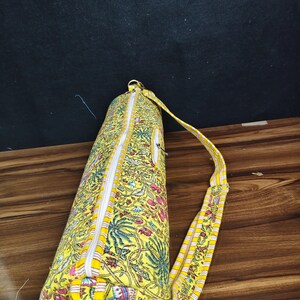 Puede incluir: Una bolsa amarilla para esterilla de yoga con un estampado floral en rosa, azul y verde. La bolsa tiene una cremallera blanca y una correa a rayas amarillas y blancas. La bolsa es cil&iacute;ndrica y parece estar hecha de tela.