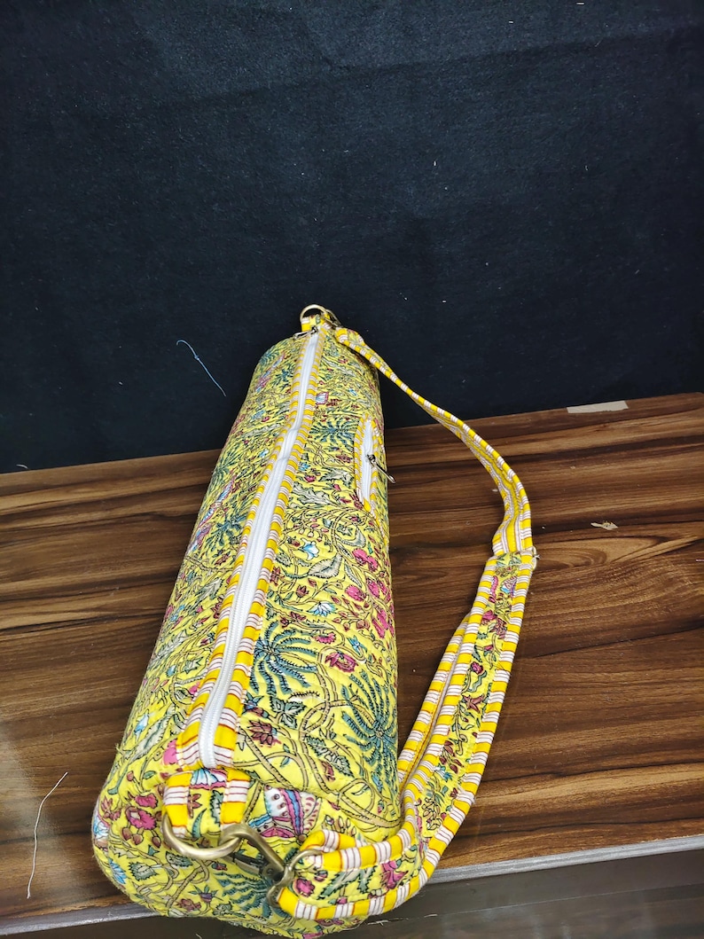 Puede incluir: Una bolsa amarilla para esterilla de yoga con un estampado floral en rosa, azul y verde. La bolsa tiene una cremallera blanca y una correa a rayas amarillas y blancas. La bolsa es cil&iacute;ndrica y parece estar hecha de tela.
