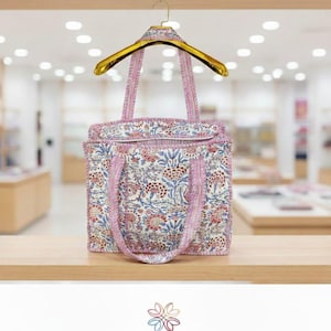 Könnte beinhalten: Eine Tragetasche mit floralem Muster und rosa Besatz und Griffen, ausgestellt auf einem Holzregal. Die Tasche weist ein sich wiederholendes Muster aus Blumen und Blättern in Blau-, Rot- und Cremetönen auf. Ein goldener Kleiderbügel stützt die Tasche.