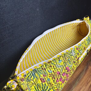 Puede incluir: Un bolso amarillo con estampado floral, cremallera blanca e interior a rayas. El bolso presenta un vibrante estampado de flores rosas y azules y hojas verdes sobre un fondo amarillo. El bolso est&aacute; abierto, revelando el interior.