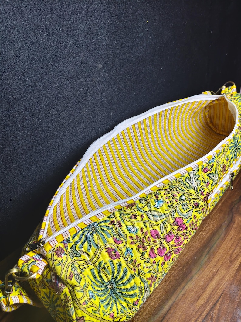 Puede incluir: Un bolso amarillo con estampado floral, cremallera blanca e interior a rayas. El bolso presenta un vibrante estampado de flores rosas y azules y hojas verdes sobre un fondo amarillo. El bolso est&aacute; abierto, revelando el interior.
