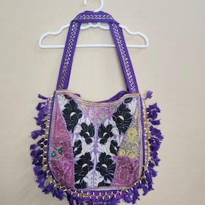 May include: A purple patchwork shoulder bag with a floral design. The bag has a gold beaded trim and purple tassels. The straps are purple with gold accents. The bag is hanging on a white hanger.