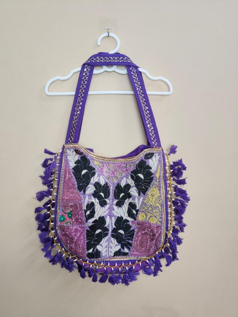Puede incluir: Bolso de hombro patchwork morado con dise&ntilde;o floral. El bolso tiene un ribete de cuentas doradas y borlas moradas. Las correas son moradas con detalles dorados. El bolso est&aacute; colgado de una percha blanca.