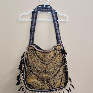 Può includere: Borsa a tracolla nera con ricami dorati intricati e motivi paisley. La borsa presenta nappe nere lungo il bordo inferiore e una tracolla nera con dettagli viola e argento. Appesa a una gruccia bianca.