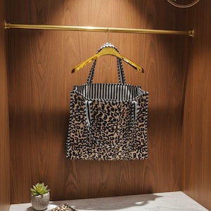 May include: A leopard print tote bag hangs on a gold hanger inside a wooden wardrobe. The bag has black and white striped lining and handles. A matching leopard print item and a small potted plant sit on a white marble shelf below.