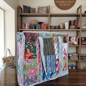 May include: A large patchwork tote bag with a variety of floral and geometric patterns in pink, green, blue, and red. The bag has a striped border and handles. The background shows other bags and the text "KHADIJA WEAVES Handcrafted Textiles".