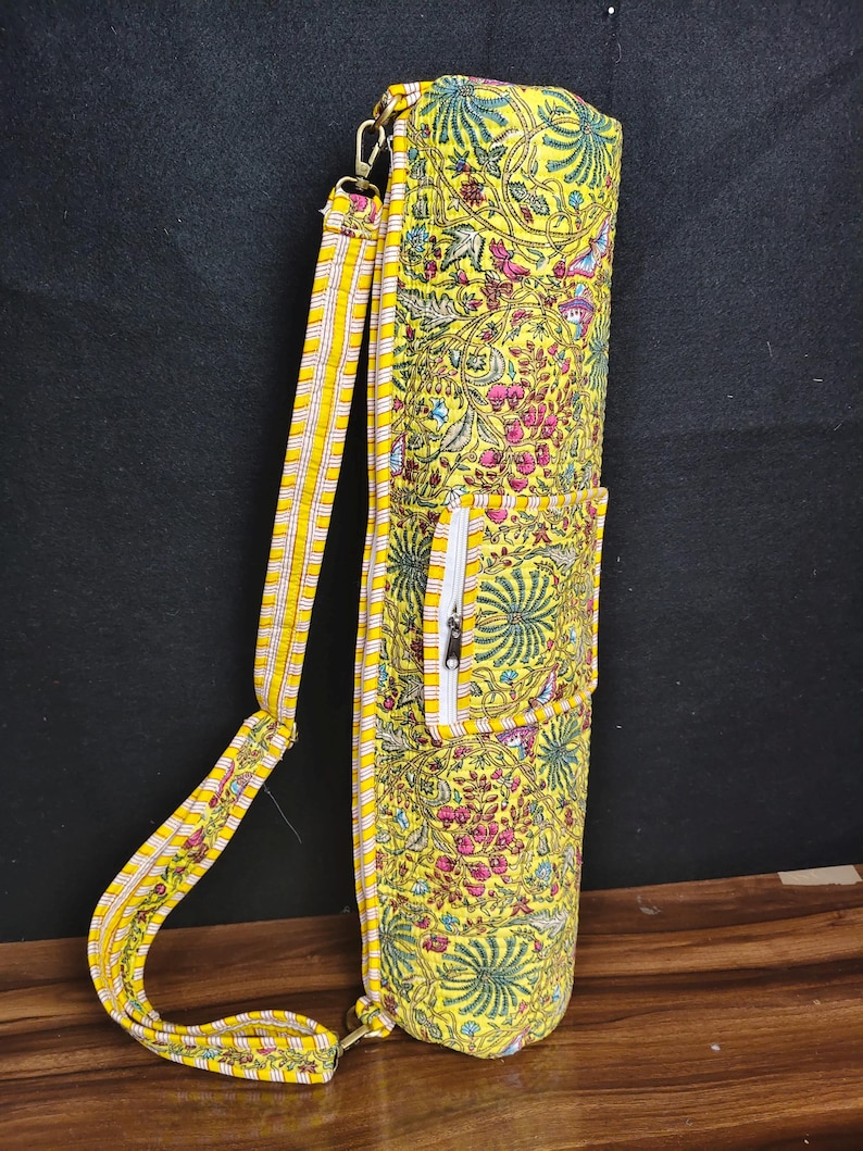 Puede incluir: Bolsa de esterilla de yoga amarilla con un estampado floral en rosa, verde y azul. La bolsa tiene un bolsillo con cremallera y una correa para el hombro a rayas. La bolsa es de tela y tiene forma cil&iacute;ndrica.