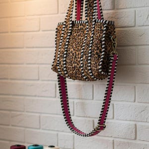 May include: A leopard print tote bag with black and white striped trim and a pink strap hangs on a wooden rack. The bag has a rectangular shape and a gold-colored clasp. The brand name "Khadija Weaves" is visible.