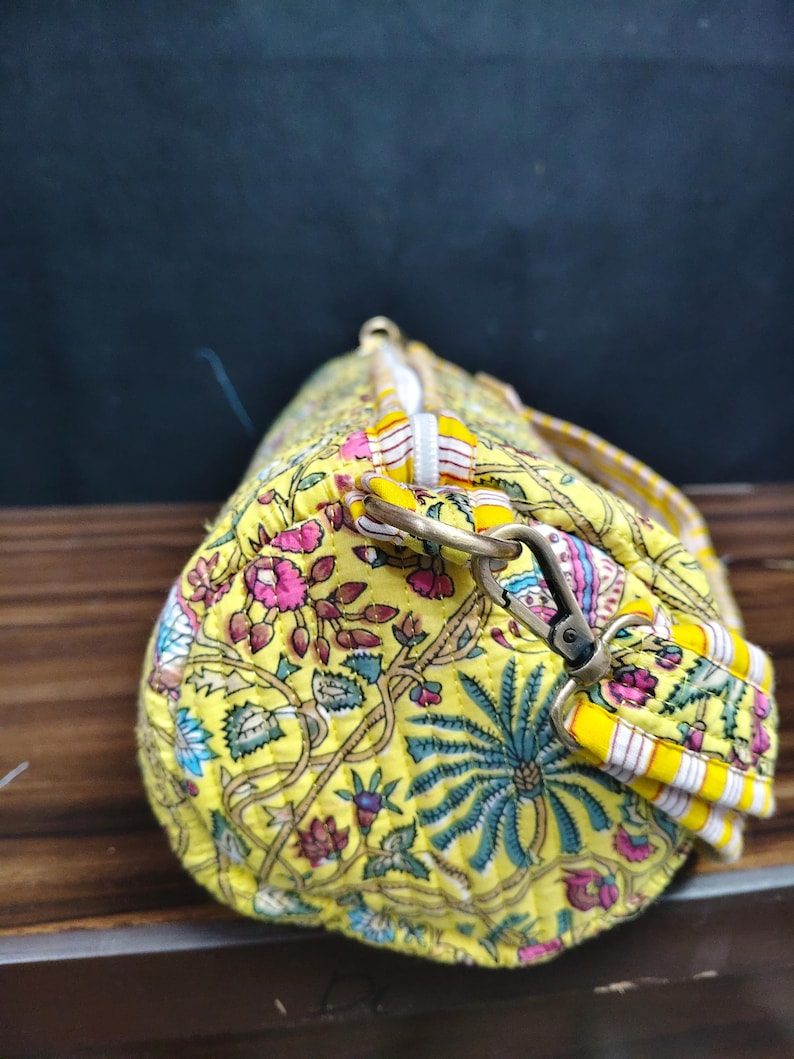 Puede incluir: Bolsa de deporte amarilla con estampado floral y dise&ntilde;o acolchado. La bolsa presenta un patr&oacute;n vibrante de flores y follaje rosas, azules y verdes sobre un fondo amarillo. Tiene una cremallera blanca y una correa para el hombro desmontable con rayas amarillas y blancas.