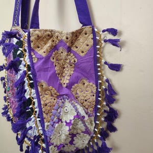 Puede incluir: Un bolso tote morado con detalles bordados en dorado y borlas moradas. El bolso presenta un dise&ntilde;o acolchado con intrincados patrones y detalles de cuentas en los bordes. El bolso tiene correas moradas.