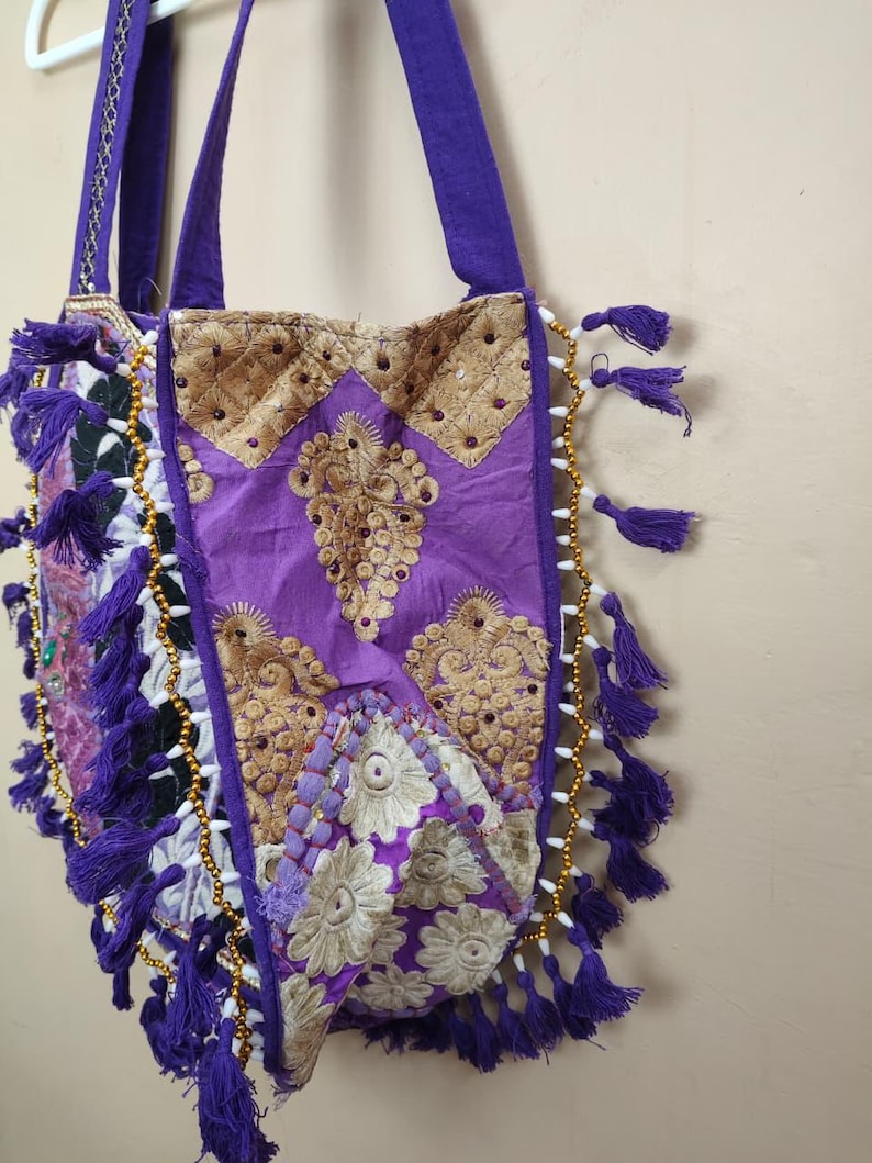 Puede incluir: Un bolso tote morado con detalles bordados en dorado y borlas moradas. El bolso presenta un dise&ntilde;o acolchado con intrincados patrones y detalles de cuentas en los bordes. El bolso tiene correas moradas.