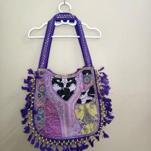 Puede incluir: Un bolso tote morado con intrincados bordados y detalles de borlas. El bolso presenta una variedad de colores, incluyendo rosa, verde y amarillo, con un ribete de cuentas doradas. Las asas y la parte superior son de color morado liso.