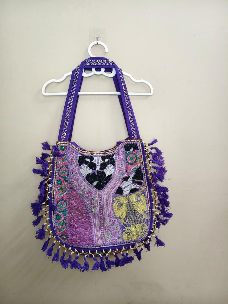 Puede incluir: Un bolso tote morado con intrincados bordados y detalles de borlas. El bolso presenta una variedad de colores, incluyendo rosa, verde y amarillo, con un ribete de cuentas doradas. Las asas y la parte superior son de color morado liso.