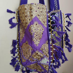 Puede incluir: Dos bolsos tote morados con intrincados bordados dorados y adornos. Los bolsos presentan borlas decorativas y detalles de cuentas en los bordes. Las asas son de color morado liso, sobre un fondo neutro.