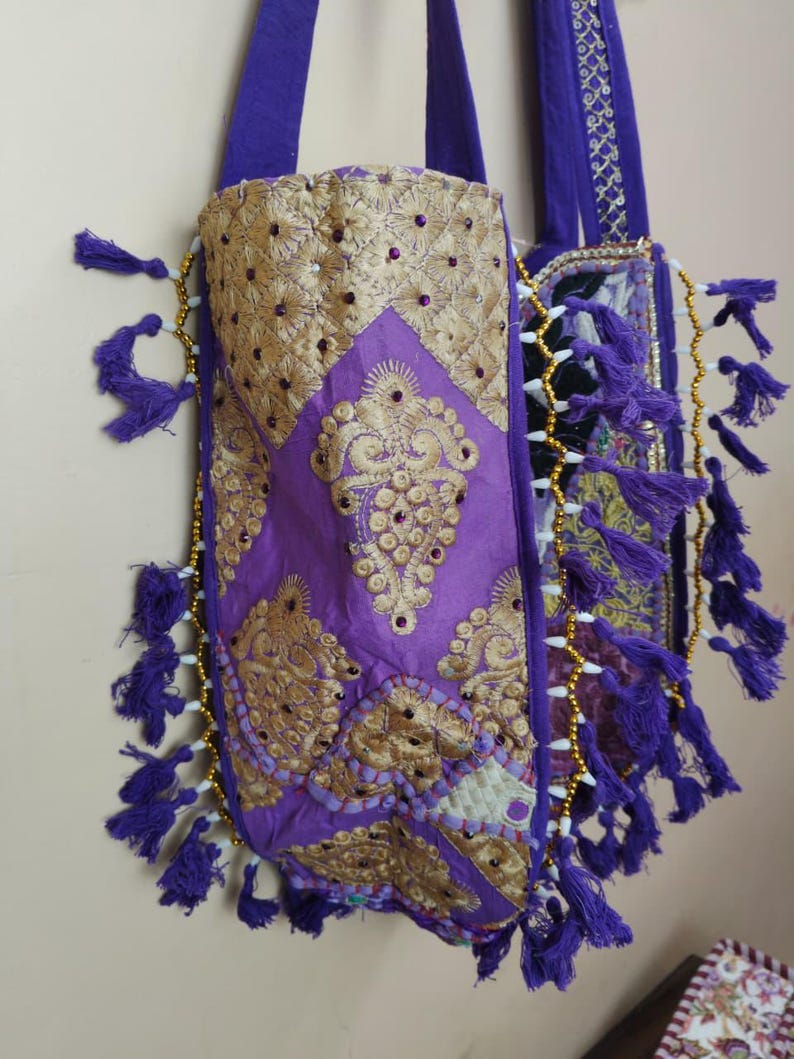 Puede incluir: Dos bolsos tote morados con intrincados bordados dorados y adornos. Los bolsos presentan borlas decorativas y detalles de cuentas en los bordes. Las asas son de color morado liso, sobre un fondo neutro.
