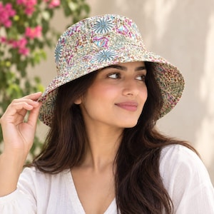 May include: A bucket hat featuring a detailed floral and botanical print in shades of blue, pink, green, and brown on a cream-coloured background. The hat has a wide, downward-sloping brim and a rounded crown, constructed from a patterned fabric.