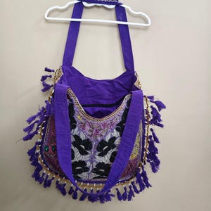 Puede incluir: Bolso de hombro morado con bordado floral y ribete de cuentas. El bolso tiene largas correas moradas y borlas moradas decorativas. Cuelga de una percha blanca contra una pared neutra.