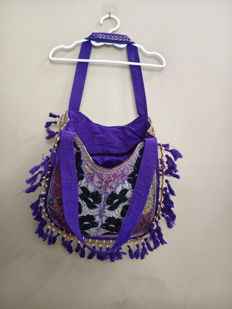 Puede incluir: Bolso de hombro morado con bordado floral y ribete de cuentas. El bolso tiene largas correas moradas y borlas moradas decorativas. Cuelga de una percha blanca contra una pared neutra.