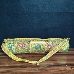 Puede incluir: Una bolsa amarilla para esterilla de yoga con un estampado floral en rosa, verde y azul. La bolsa tiene un bolsillo con cremallera y una correa amarilla a juego. La bolsa mide aproximadamente 71 cm de largo.