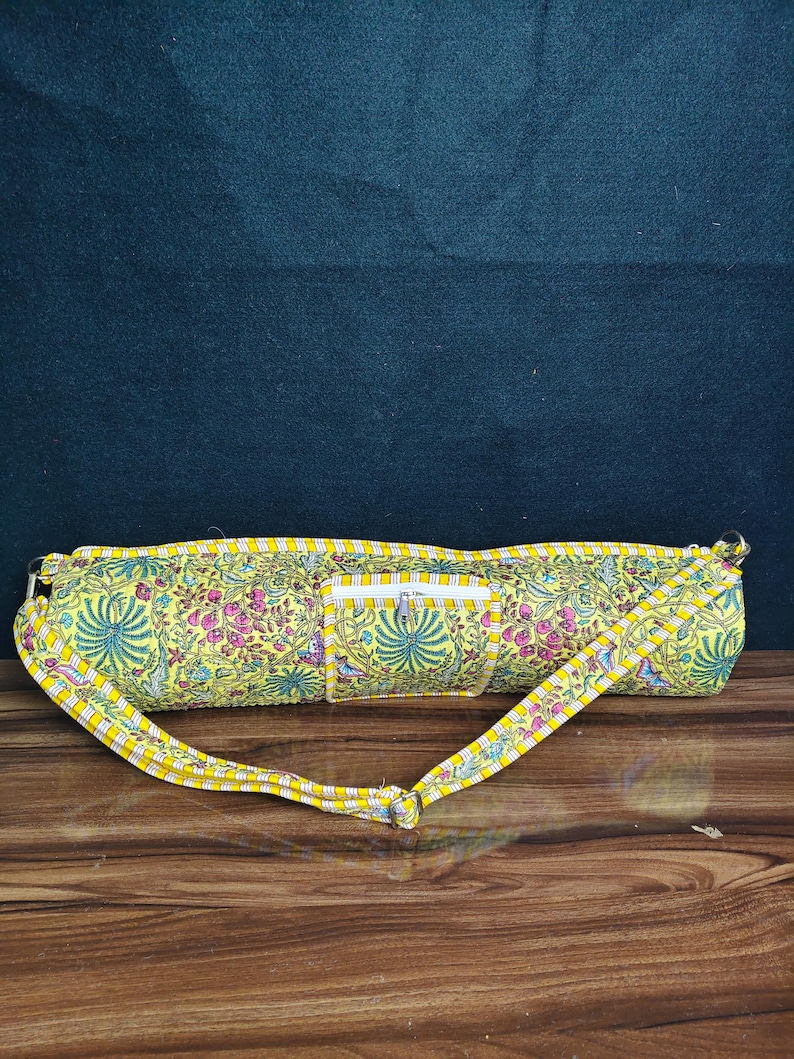 Puede incluir: Una bolsa amarilla para esterilla de yoga con un estampado floral en rosa, verde y azul. La bolsa tiene un bolsillo con cremallera y una correa amarilla a juego. La bolsa mide aproximadamente 71 cm de largo.