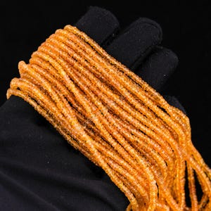 May include: A collection of strands of small, round, orange beads. The beads are tightly strung together, creating a textured surface. The beads are held against a black background, highlighting their color and detail. The beads appear to be made of a translucent material.