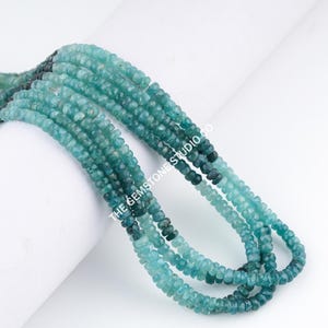 Könnte beinhalten: Stränge facettierter, teal-farbener Edelsteinperlen. Die Perlen wechseln von einem dunkleren Teal zu einem helleren Farbton und erzeugen einen Ombre-Effekt. Auf dem Bild steht "The Gemstone Studio Co.".