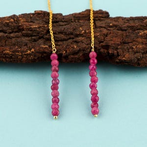 May include: A pair of threader earrings featuring thin gold chains, each ending in a vertical strand of small, faceted, round, deep pink ruby beads. The beads are arranged in a single column, creating a delicate and elongated drop design.