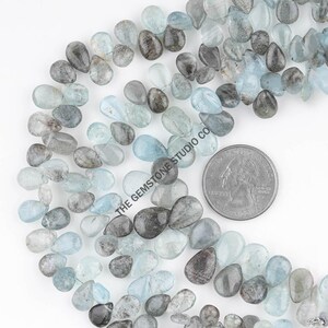 May include: A close-up of a collection of aquamarine gemstone beads in various shades of blue and gray. The beads are teardrop-shaped and strung together. A US quarter is included for size reference. The text "THE GEMSTONE STUDIO CO" is visible.